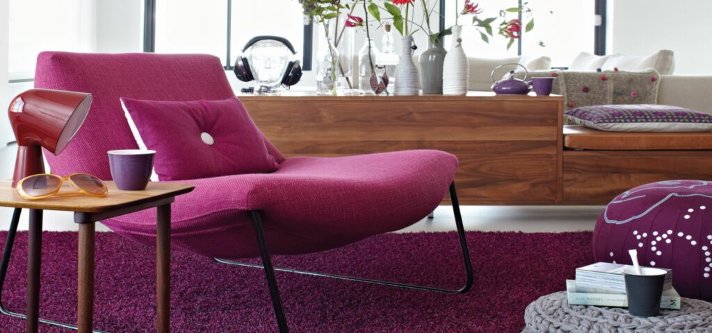 Ein stilvolles Wohnzimmer mit einem Sessel in pinknem Polsterstoff, auf einem passenden Teppich. Dekoriert mit Kissen in ausgewählten Möbelstoffen.
