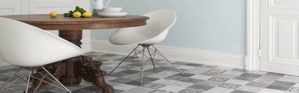 Bild Produkte: Ein Esszimmer mit einem runden Holztisch mit verzierten Beinen, zwei modernen weißen Stühlen, vor einer hellblauen Wand und CV Belag in Betonfliesenoptik.
