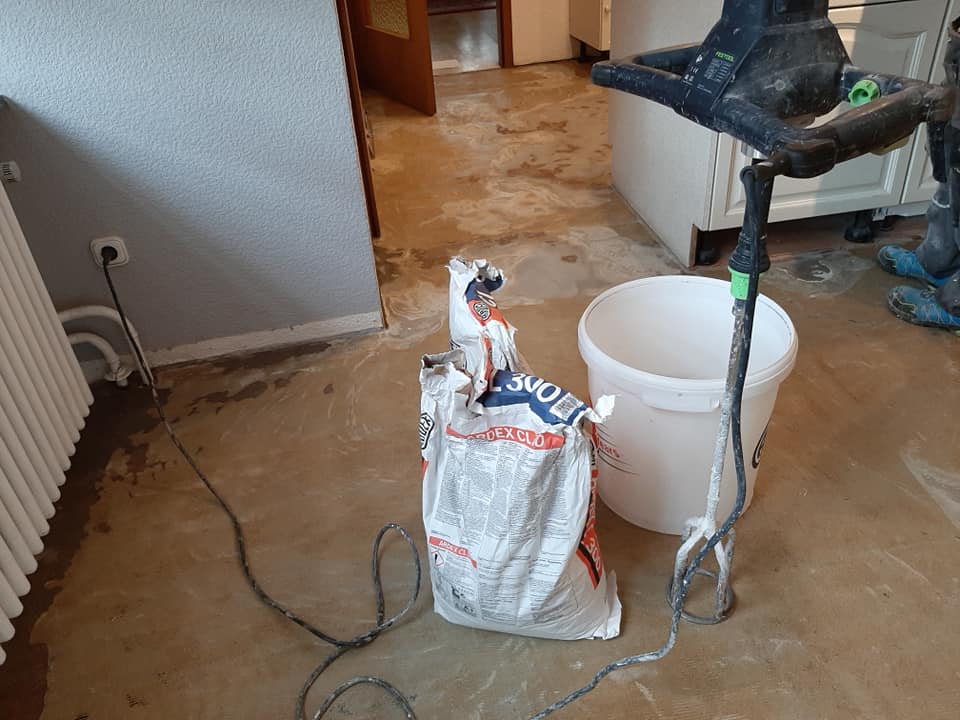 Renovierungswerkzeuge, darunter ein Sack Spachtelmasse, ein weißer Eimer und eine Rührbohrmaschine in einem Raum unter Bodenverlegung.